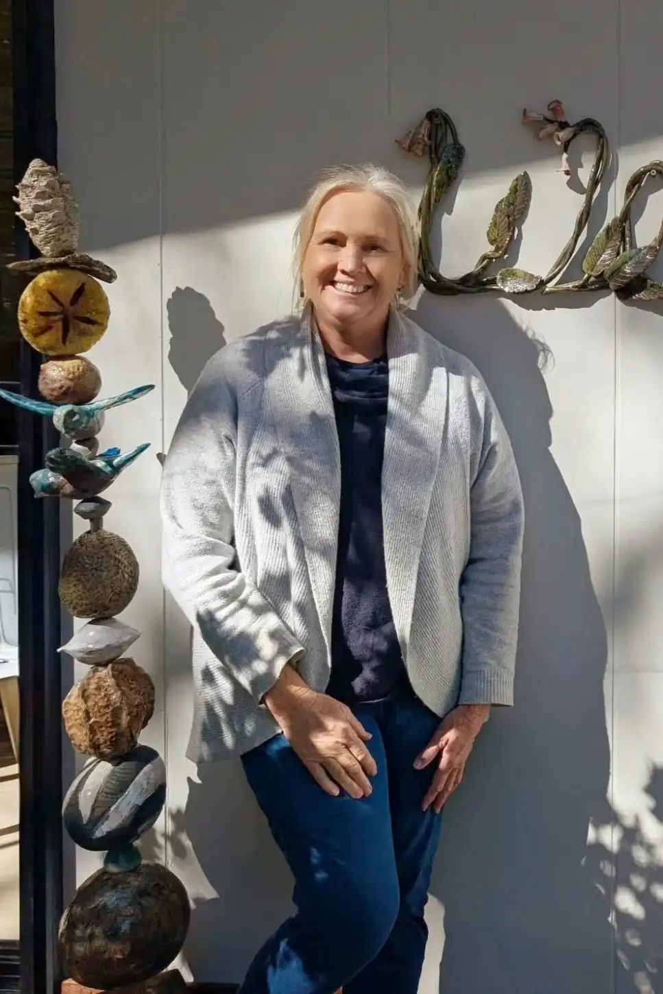 Deb Howells smiling beside a ceramic totem and a clay “Welcome” sign on her studio wall
